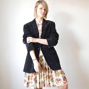 May include: A black corduroy blazer over a floral print dress. The blazer has a classic notched lapel and button closure. The dress features a cream background with a pattern of red, yellow, and green flowers.