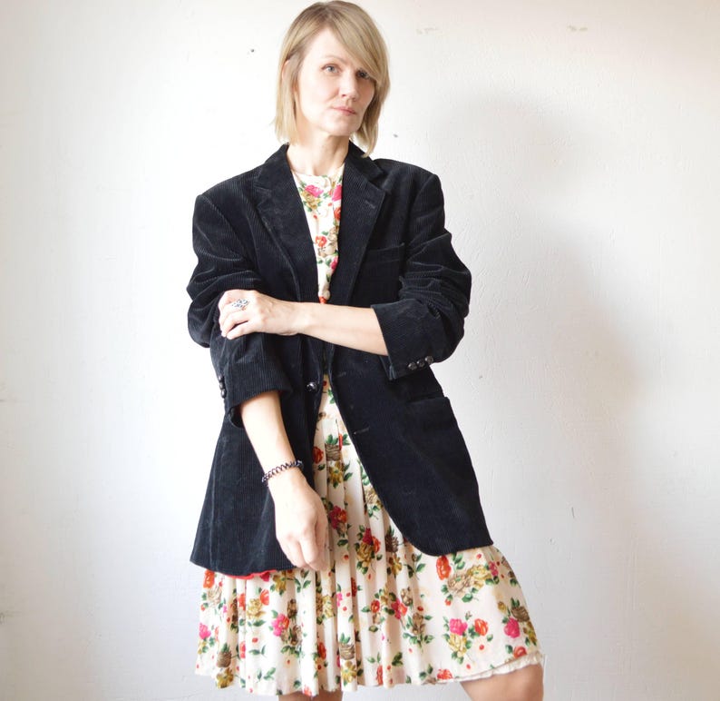 May include: A black corduroy blazer over a floral print dress. The blazer has a classic notched lapel and button closure. The dress features a cream background with a pattern of red, yellow, and green flowers.