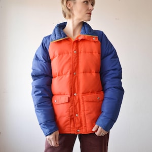 Könnte beinhalten: Eine Vintage-Steppjacke mit einem leuchtend orangenen Oberkörper und blauen Ärmeln. Die Jacke hat einen gelben Reißverschluss und einen Druckknopfverschluss.