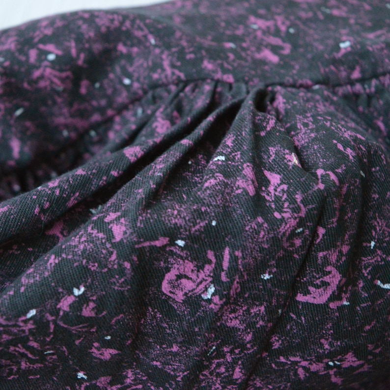 May include: Close-up of a black fabric with a purple and white speckled pattern. The fabric is gathered and has a soft, textured feel.