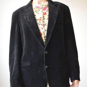 May include: A black corduroy blazer with a two-button closure and notched lapels. The blazer has a classic design and a textured appearance. A floral patterned shirt is visible underneath the blazer.