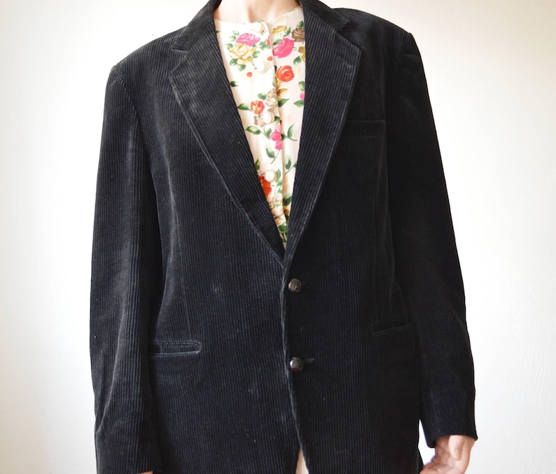 May include: A black corduroy blazer with a two-button closure and notched lapels. The blazer has a classic design and a textured appearance. A floral patterned shirt is visible underneath the blazer.