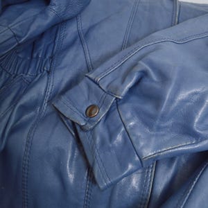 May include: Close-up of a blue leather jacket. The jacket features a buttoned cuff and detailed stitching. The leather has a slightly worn appearance, with visible creases and subtle color variations.