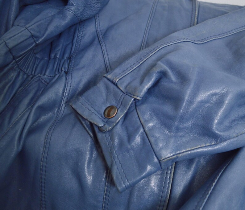 May include: Close-up of a blue leather jacket. The jacket features a buttoned cuff and detailed stitching. The leather has a slightly worn appearance, with visible creases and subtle color variations.
