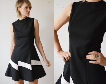 May include: A sleeveless black dress with a flared skirt and white geometric accents. The dress has a high neckline and a fitted bodice. The white accents are angled and create a bold, graphic design. The dress is made of a smooth, solid fabric.