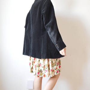 May include: A black corduroy blazer with three buttons on the sleeves. The blazer is worn over a floral print skirt. The skirt has a cream background with red, pink, and yellow flowers.