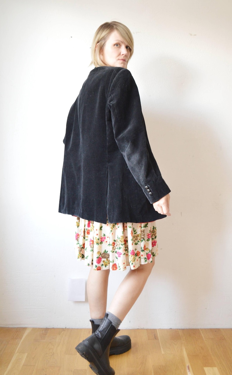 May include: A black corduroy blazer with three buttons on the sleeves. The blazer is worn over a floral print skirt. The skirt has a cream background with red, pink, and yellow flowers.