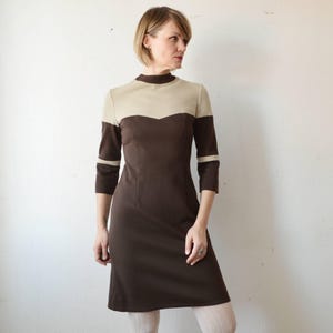 Puede incluir: Vestido vintage marrón y beige con cuello alto y mangas tres cuartos. El vestido presenta un diseño de bloques de color con beige en el escote y la parte superior del pecho, y un cuerpo marrón. Las mangas tienen rayas beige cerca del codo. El vestido es hasta la rodilla.