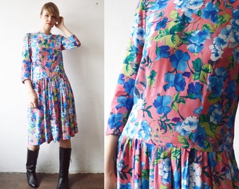Könnte beinhalten: Ein rosa-blaues Blumenkleid mit langen Ärmeln und einem gerafften Rock. Das Kleid hat ein figurbetontes Oberteil und einen leicht ausgestellten Rock.