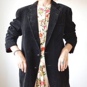 May include: A black corduroy blazer with a two-button closure. The blazer has a classic notched lapel and a tailored fit. The sleeves are long, and the jacket is worn over a floral print dress. The blazer is a versatile piece for any wardrobe.