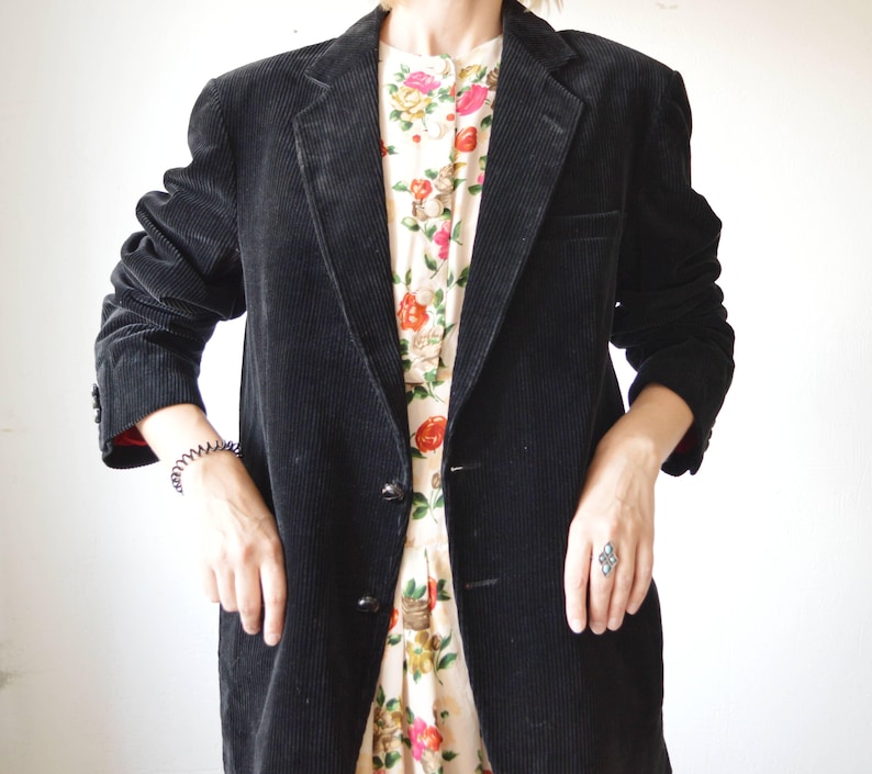 May include: A black corduroy blazer with a two-button closure. The blazer has a classic notched lapel and a tailored fit. The sleeves are long, and the jacket is worn over a floral print dress. The blazer is a versatile piece for any wardrobe.