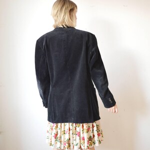 May include: A dark navy corduroy blazer with a two-button closure and a notched lapel. The blazer is worn over a floral print skirt with a cream background and pink and red flowers. The blazer has a classic, tailored fit.