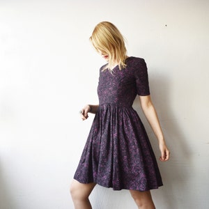 May include: A woman wearing a black and purple floral print dress with a short sleeve and a fit and flare silhouette.