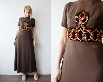 May include: A brown maxi dress with a beaded embellishment around the waist. The embellishment is made of gold and orange beads and has a floral design.