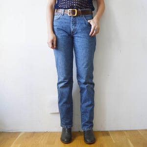 May include: A person wearing a pair of blue denim jeans with a brown leather belt and a gold buckle. The person is wearing black ankle boots.