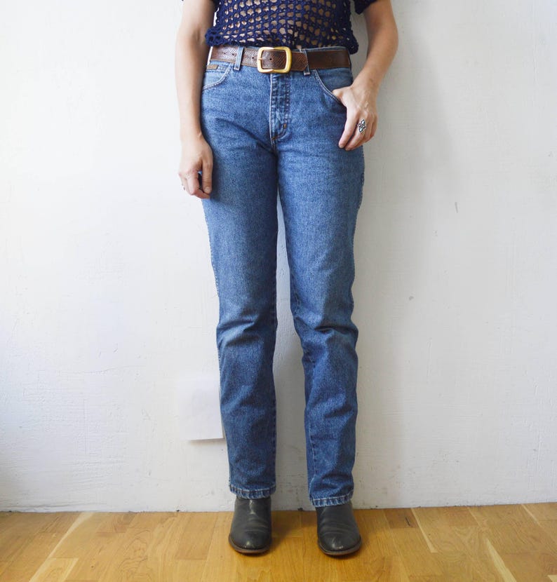 May include: A person wearing a pair of blue denim jeans with a brown leather belt and a gold buckle. The person is wearing black ankle boots.