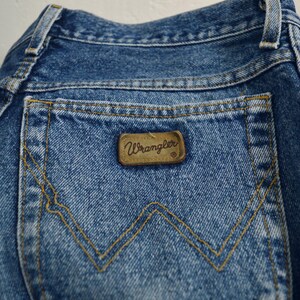 May include: A close-up of the back pocket of a pair of blue denim jeans. The pocket has a brown leather patch with the word "Wrangler" printed on it.