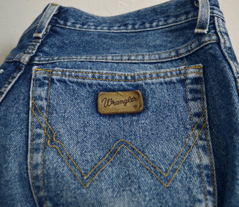 May include: A close-up of the back pocket of a pair of blue denim jeans. The pocket has a brown leather patch with the word "Wrangler" printed on it.