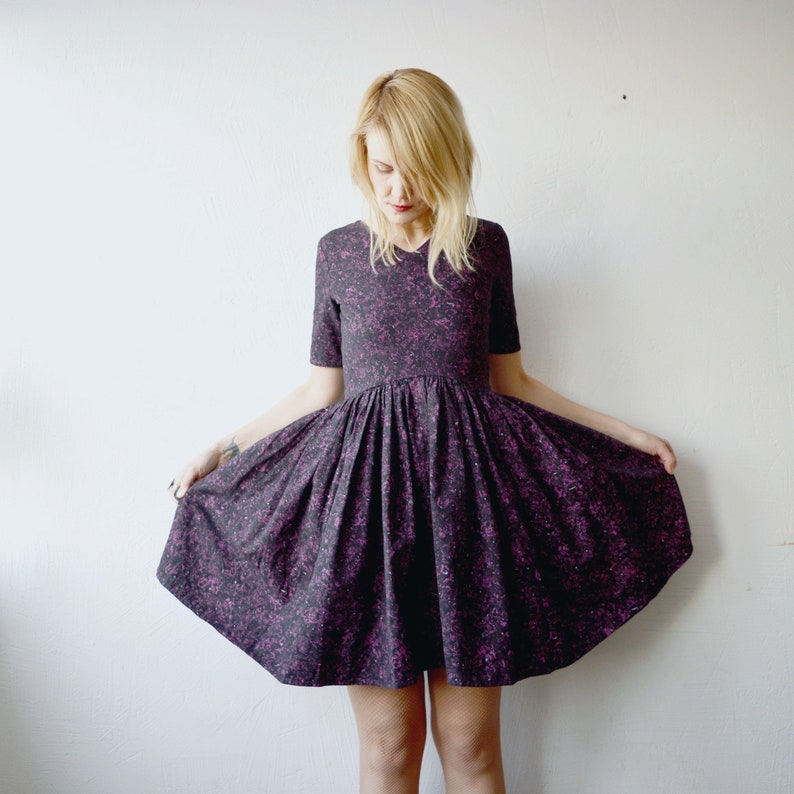 May include: A woman wearing a short, dark purple dress with a pink floral pattern. The dress has a gathered skirt and short sleeves.