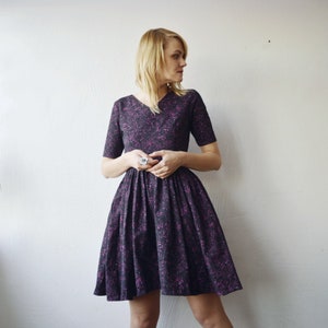 May include: A woman wearing a purple and black short-sleeved dress with a v-neckline. The dress has a scattered pattern of small pink and white dots.