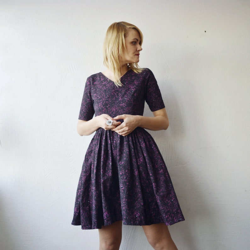 May include: A woman wearing a purple and black short-sleeved dress with a v-neckline. The dress has a scattered pattern of small pink and white dots.