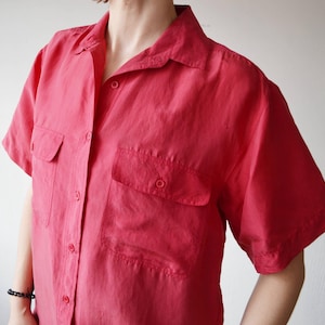 Puede incluir: Una camisa roja de manga corta con dos bolsillos delanteros y cuello. La camisa tiene cierre de botones y está hecha de una tela ligera. Las mangas son cortas y la camisa tiene un corte relajado.