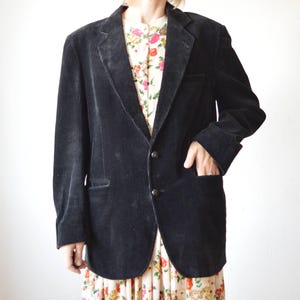 May include: A black corduroy blazer with a two-button closure and a notched lapel. The blazer has two front pockets and a pocket on the chest. The blazer is worn over a floral print dress.
