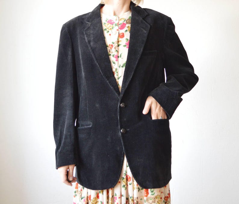 May include: A black corduroy blazer with a two-button closure and a notched lapel. The blazer has two front pockets and a pocket on the chest. The blazer is worn over a floral print dress.