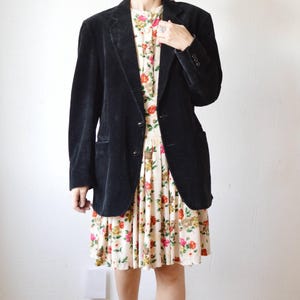 May include: A black corduroy blazer over a floral print dress. The dress features a cream background with red, orange, and yellow flowers. The outfit is completed with black boots and gray socks. The blazer has two buttons and a notched lapel.