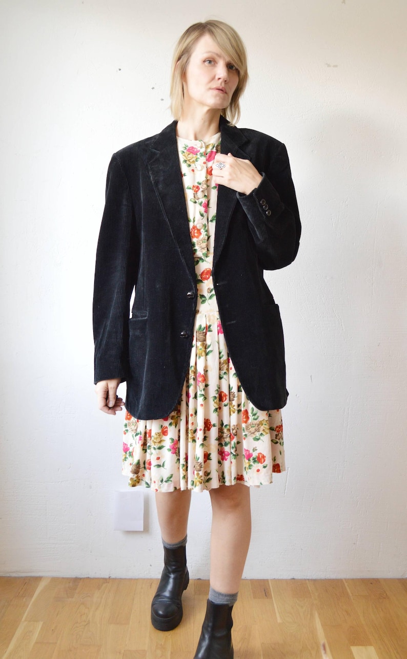 May include: A black corduroy blazer over a floral print dress. The dress features a cream background with red, orange, and yellow flowers. The outfit is completed with black boots and gray socks. The blazer has two buttons and a notched lapel.