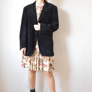 May include: A black corduroy blazer over a floral print dress. The dress has a cream background with red, pink, and yellow flowers. Black platform boots complete the outfit. The blazer has a classic notched lapel.