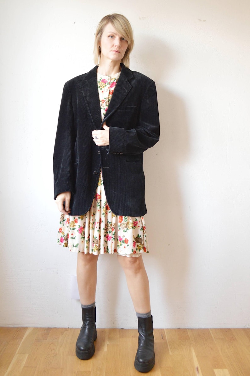 May include: A black corduroy blazer over a floral print dress. The dress has a cream background with red, pink, and yellow flowers. Black platform boots complete the outfit. The blazer has a classic notched lapel.