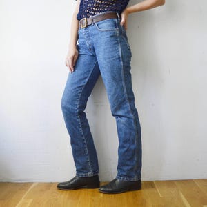 May include: A pair of blue denim jeans with a straight leg fit. The jeans have a brown leather belt and are being worn by a person standing on a wooden floor.