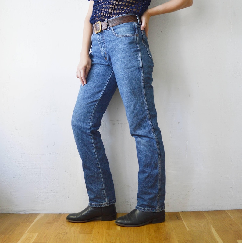 May include: A pair of blue denim jeans with a straight leg fit. The jeans have a brown leather belt and are being worn by a person standing on a wooden floor.