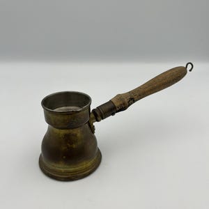 May include: A small, antique brass coffee pot with a wooden handle. The pot has a rounded base and a narrow neck, with a handle that curves upwards and ends in a hook. The brass has a tarnished, aged appearance.