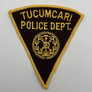 Puede incluir: Parche triangular con las palabras "TUCUMCARI POLICE DEPT." bordadas en dorado sobre fondo negro. El parche tiene un borde dorado y un emblema dorado en el centro.