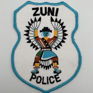 May include: Embroidered patch with a shield shape and a turquoise border. The patch features the words "ZUNI POLICE" in black text. A Native American figure with outstretched arms is in the center, wearing traditional clothing.
