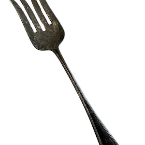 Puede incluir: Un tenedor de plata vintage con cuatro púas. El tenedor tiene un mango largo y delgado con detalles decorativos en la base. El metal tiene un aspecto ligeramente deslustrado, lo que sugiere edad y uso. El tenedor está aislado sobre un fondo blanco.