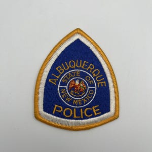 Puede incluir: Un parche bordado en forma de escudo con fondo azul, borde dorado y ribete interior blanco. Presenta el texto ALBUQUERQUE POLICE en dorado, rodeando un emblema circular central con el texto STATE OF NEW MEXICO y un gráfico de un ave.