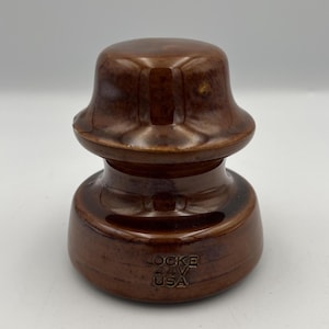 May include: A brown ceramic insulator with a glossy finish. The insulator has a rounded top and a wider base, with the text "LOCKE" and "USA" embossed on the side. This vintage piece is likely from the electrical industry.