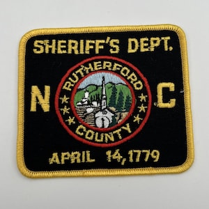 Puede incluir: Un parche bordado en negro y dorado con el texto "SHERIFF'S DEPT." y "RUTHERFORD COUNTY" que rodea un emblema circular con una iglesia y árboles. El parche también incluye el texto "N C" y "APRIL 14, 1779".