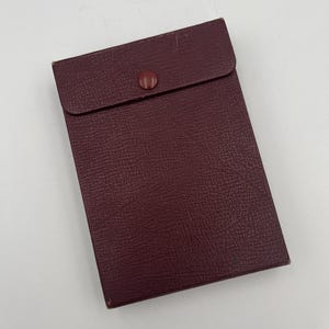 Może przedstawiać: Prostokątne etui w kolorze bordowym, z teksturowaną powierzchnią i klapką zabezpieczoną pasującym guzikiem. Etui, prawdopodobnie wykonane z materiału skóropodobnego, jest zamknięte.