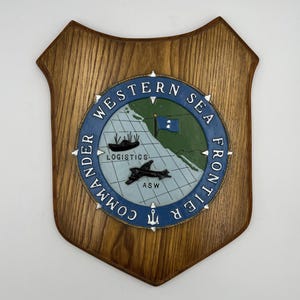 May include: Wooden plaque with a shield shape. The plaque features a circular emblem with the text "WESTERN SEA FRONTIER COMMANDER" in blue. The emblem includes a map, a ship, a plane, and the word "LOGISTICS" and "ASW".