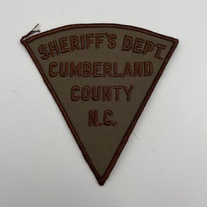 May include: A triangular patch with the text "SHERIFF'S DEPT CUMBERLAND COUNTY N.C." in brown embroidery on a green background. The patch has a brown border and a small brown thread at the top.