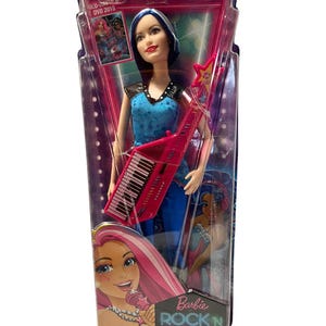 Puede incluir: Muñeca Barbie Rock 'n Royals con cabello azul y un teclado rosa. La muñeca lleva un atuendo azul y negro con detalles plateados. El empaque presenta el logotipo de Barbie Rock 'n Royals y una imagen de la muñeca cantando.