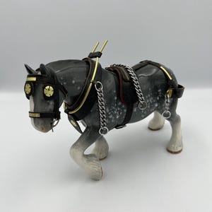 Puede incluir: Figura de caballo de cerámica en posición de pie, con un arnés detallado con detalles dorados y una cadena plateada. El caballo es principalmente gris con motas blancas, y sus cascos son blancos. El arnés es negro con detalles dorados.