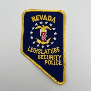 May include: A dark blue embroidered patch shaped like the state of Nevada with a yellow border. It features the text NEVADA LEGISLATURE SECURITY POLICE in yellow, a central emblem with a yellow eagle, and a circle of white stars on a blue field.