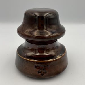 Puede incluir: Un aislador eléctrico de cerámica marrón con una parte superior e inferior redondeadas. El aislador tiene un acabado brillante y el texto "LOCKE USA" es visible en el lateral. Esta pieza vintage es probablemente de principios o mediados del siglo XX.