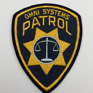 May include: A navy blue and gold patch with the words "OMNI SYSTEMS PATROL" in gold lettering. The patch features a gold star with a white scale in the center. The patch has a navy blue background and a gold border.