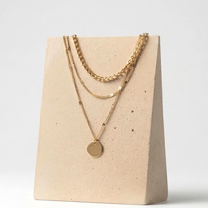 Puede incluir: Un collar de capas en tono dorado con tres cadenas de estilos diferentes. La cadena superior es de eslabones clásicos, la del medio es una cadena fina y la inferior presenta un colgante circular, plano y liso en la parte central.